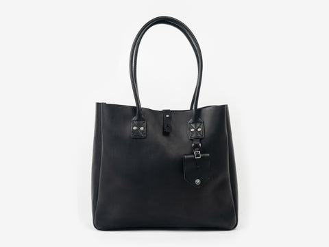 Front view of a medium-sized black leather tote with handles for your shoulder. The tote features a removable luggage tag below one of the handles. The bag is set against a neutral background.