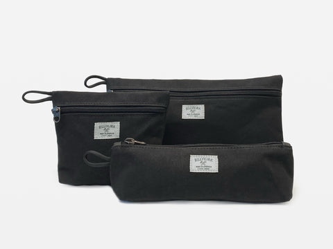 Three different sized black waxed canvas pouches with zipper closures arranged together on a neutral background.