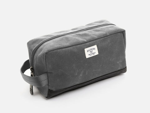 A rectangular ash grey waxed canvas toiletry bag with a zipper, a grey nylon webbing handle, a black leather base, and a off-white Billykirk brand label on the front.
