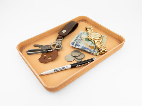 A tan leather tray holds a keychain with keys, a brown leather fob, a gold wristwatch, folded cash, coins, and a white and black pen with BILLYKIRK written on it, all on a white background.