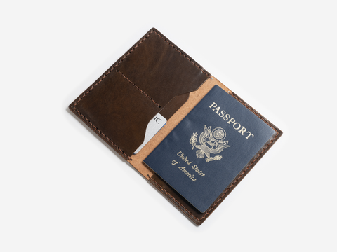 A brown leather wallet is open, displaying a blue United States passport and a partially visible white card inside. The wallet and its contents are set against a plain white background.