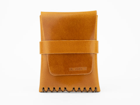 A tan leather wallet with a front flap and a horizontal strap, featuring visible black stitching at the bottom and the word BILLYKIRK embossed on the front. The wallet is photographed against a white background.