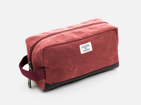 A rectangular nantucket red waxed canvas toiletry bag with a zipper, a burgundy nylon webbing handle, a black leather base, and an off-white Billykirk brand label on the front.