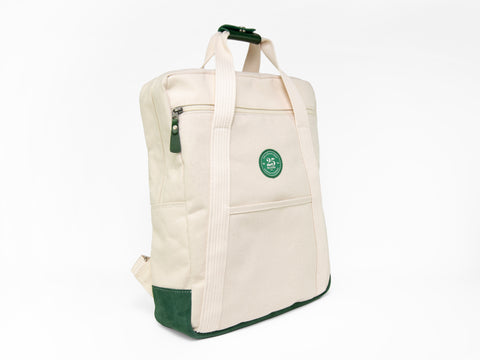 3/4 view of a natural canvas backpack tote with matching cotton straps, green suede bottom, and a green 25th anniversary patch sewn on the front. The bag positioned upright against a white background.