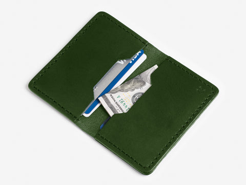 A green leather wallet is open, revealing a blue credit card, a white card, and a folded U.S. hundred dollar bill inside two pockets against a white background.