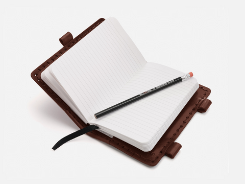 An open lined notebook with a brown leather cover and a black pencil resting on its pages. A black ribbon bookmark is visible, and the notebook is set against a white background.