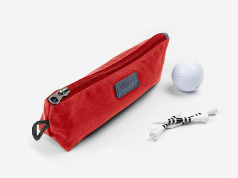 A red zippered pouch lies next to a white golf ball and three white golf tees with black markings, all on a white background.