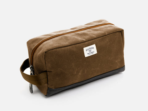 A rectangular brush brown waxed canvas toiletry bag with a zipper, a brush brown nylon webbing handle, a black leather base, and an off-white Billykirk brand label on the front.