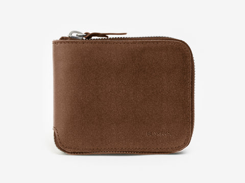 A brown suede zippered wallet with rounded edges and a visible metal zipper, placed on a plain white background. The brand name is subtly embossed in the lower right corner.