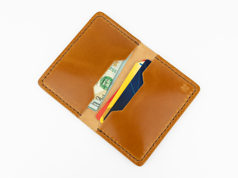 A tan cordovan leather wallet open to reveal paper currency and two credit cards inside, photographed on a white background.
