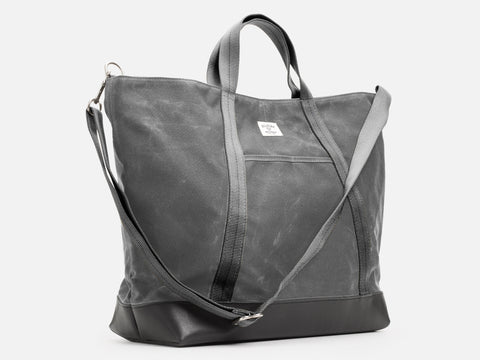 A 3/4 view of an ash waxed canvas tote bag with 1" nylon ash grey handles and shoulder strap, a small off-white label on the front, and a black leather bottom, set against a plain white background.