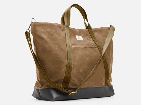 A 3/4 view of a brush brown waxed canvas tote bag with 1" nylon brush brown handles and shoulder strap, a small off-white label on the front, and a black leather bottom, set against a plain white background.