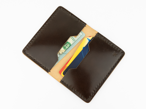 A dark brown cordovan leather wallet open to reveal paper currency and two credit cards inside, photographed on a white background.