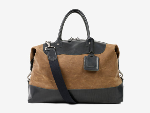 A brown and black leather travel duffel bag with black handles, a shoulder strap, and a luggage tag attached, set against a plain light background.