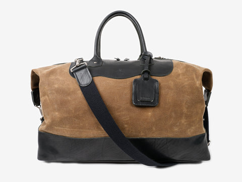 Brown and black leather duffel bag with a top handle, detachable shoulder strap, and luggage tag, photographed on a plain white background.