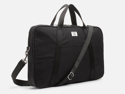 A black rectangular laptop bag with a top handle, front pocket, detachable shoulder strap, and a small white label on the front, against a plain white background.