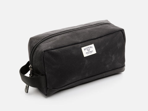 A rectangular black waxed canvas toiletry bag with a zipper, a black nylon webbing handle, a black leather base, and an off-white Billykirk brand label on the front.