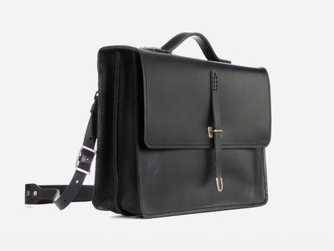 Black leather satchel with a top handle, adjustable shoulder strap, and a front flap secured by a buckle. The design is sleek and minimal, set against a plain white background.