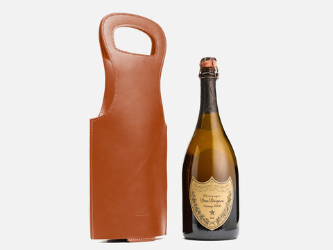 A bottle of Dom Pérignon champagne stands next to a brown leather wine carrier with a handle, both set against a plain white background.
