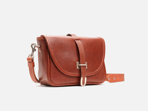 A small, brown leather crossbody bag with a textured finish, flap closure, silver buckle, and adjustable strap, displayed against a plain white background.