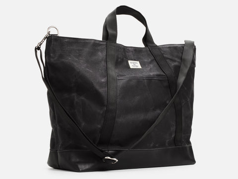 A 3/4 view of a black waxed canvas tote bag with 1" nylon black handles and shoulder strap, a small off-white label on the front, and a black leather bottom, set against a plain white background.