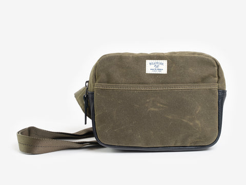 A small olive waxed canvas crossbody bag with a front pocket, adjustable strap, and a cream colored rectangular Billykirk brand patch stitched on the front.