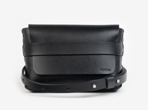 A black leather belt crossbody bag with a smooth finish, featuring a flap closure, subtle embossed branding, and an adjustable strap with silver-tone collar buttons, displayed against a white background.