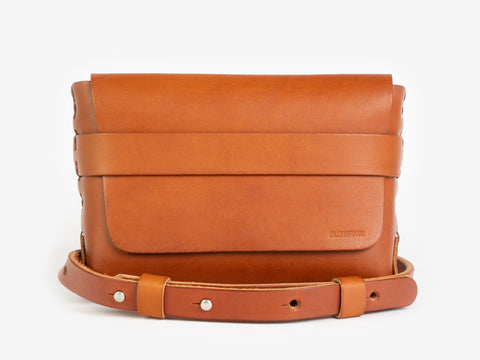 A tan leather belt crossbody bag with a smooth finish, featuring a flap closure, subtle embossed branding, and an adjustable strap with silver-tone collar buttons, displayed against a white background.