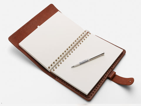 An open brown leather-bound notebook with blank white pages and a spiral binding lies on a white surface. A silver pen is resting diagonally across the pages.