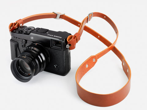 A black Fujifilm X-Pro2 camera with a lens attached, shown with a brown leather neck strap, placed on a white surface.