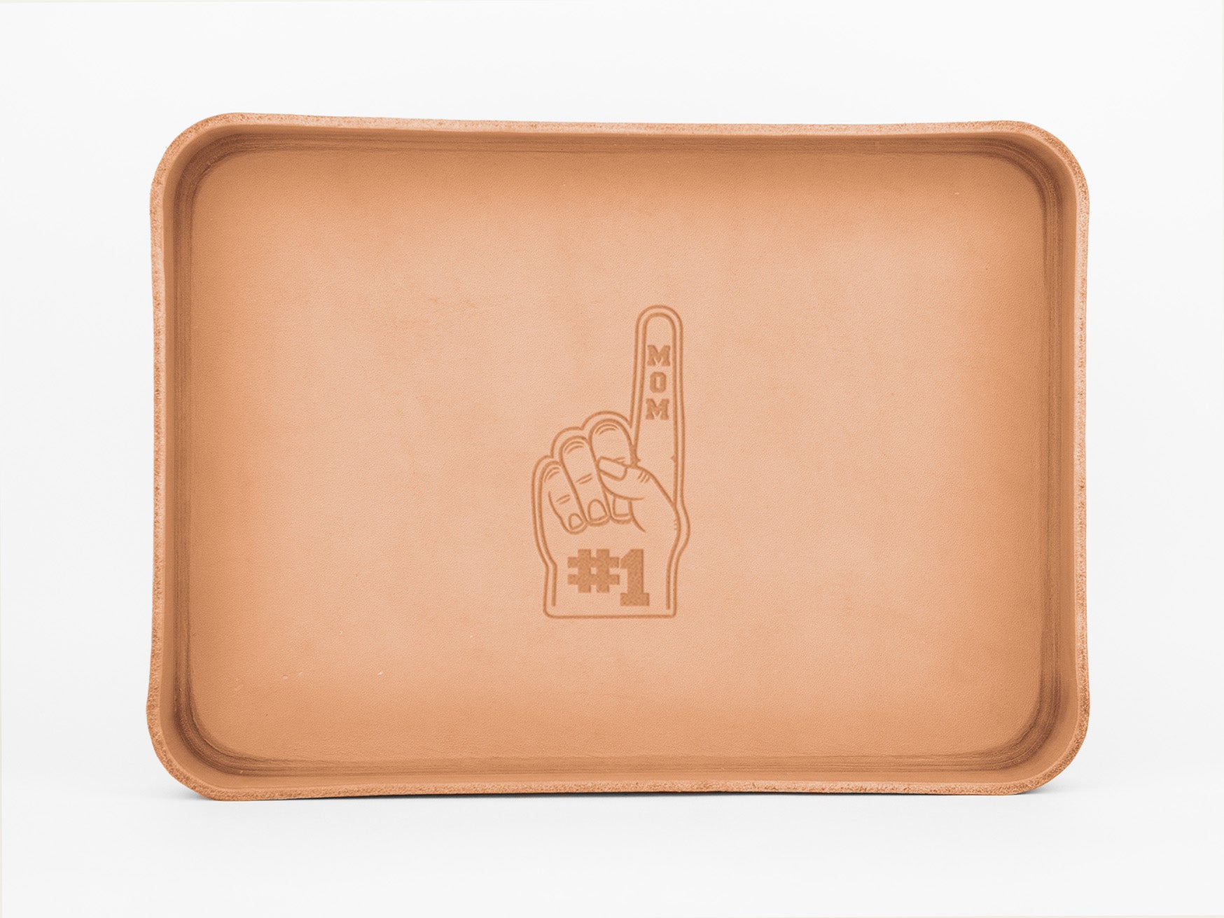 A rectangular tan leather tray features an embossed design of a foam finger hand with the raised index finger and the words MOM #1 on it. The background is plain white.