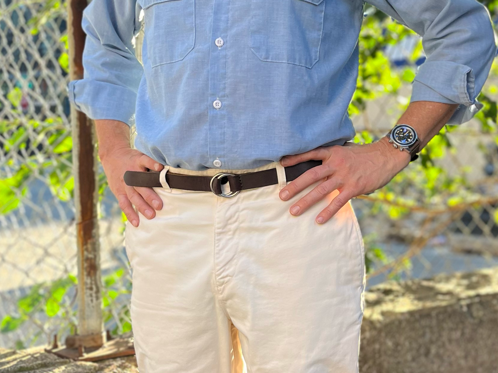 A person stands outdoors with hands on hips, wearing a light blue button-up shirt, beige pants, a brown belt, and a wristwatch. A chain-link fence and greenery are visible in the background.
