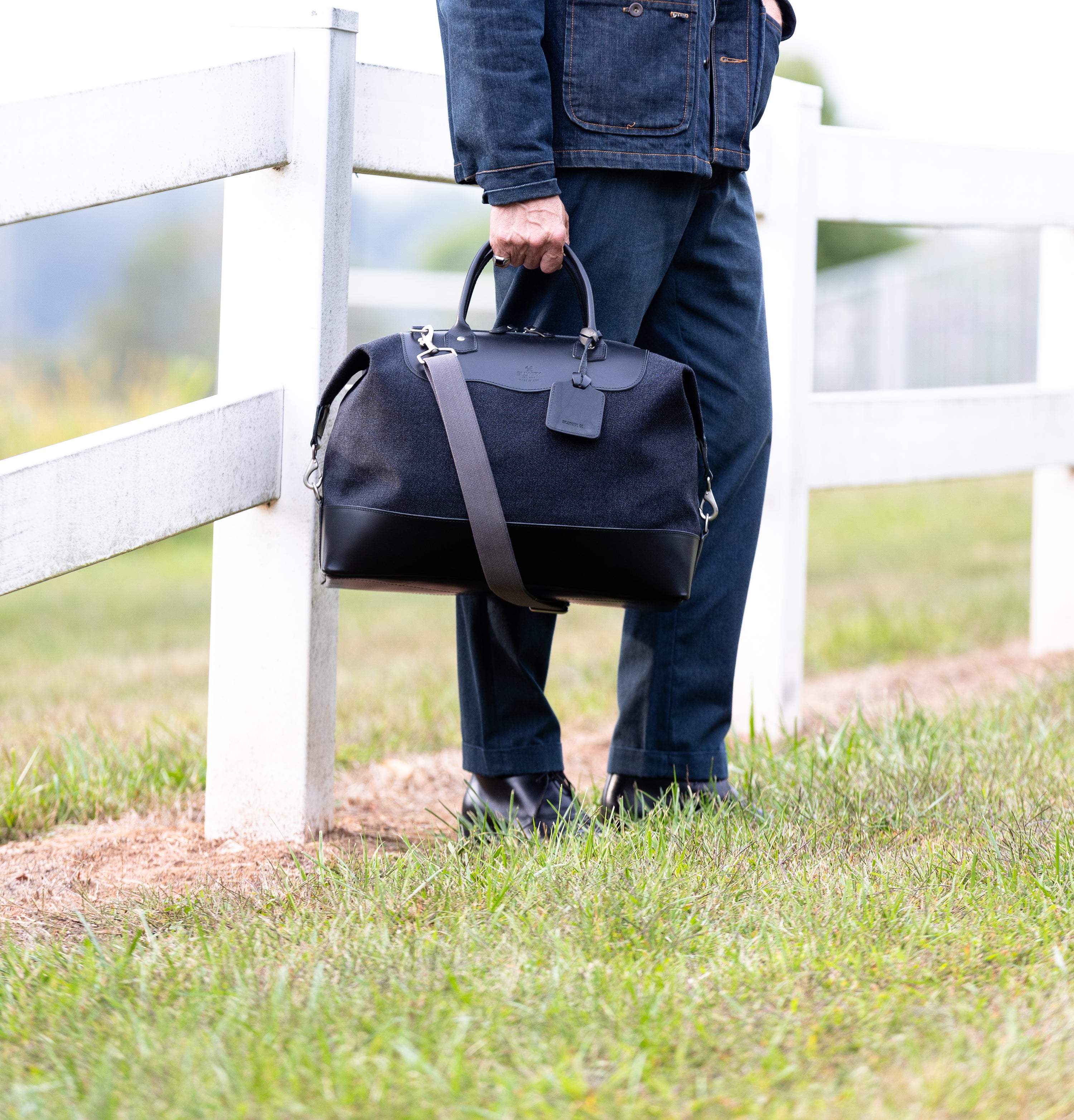 Person holding a black carryall bag outdoors near a white fence