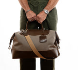 Man holding a taupe and olive leather carryall bag with beige cotton webbing shoulder strap on a white background