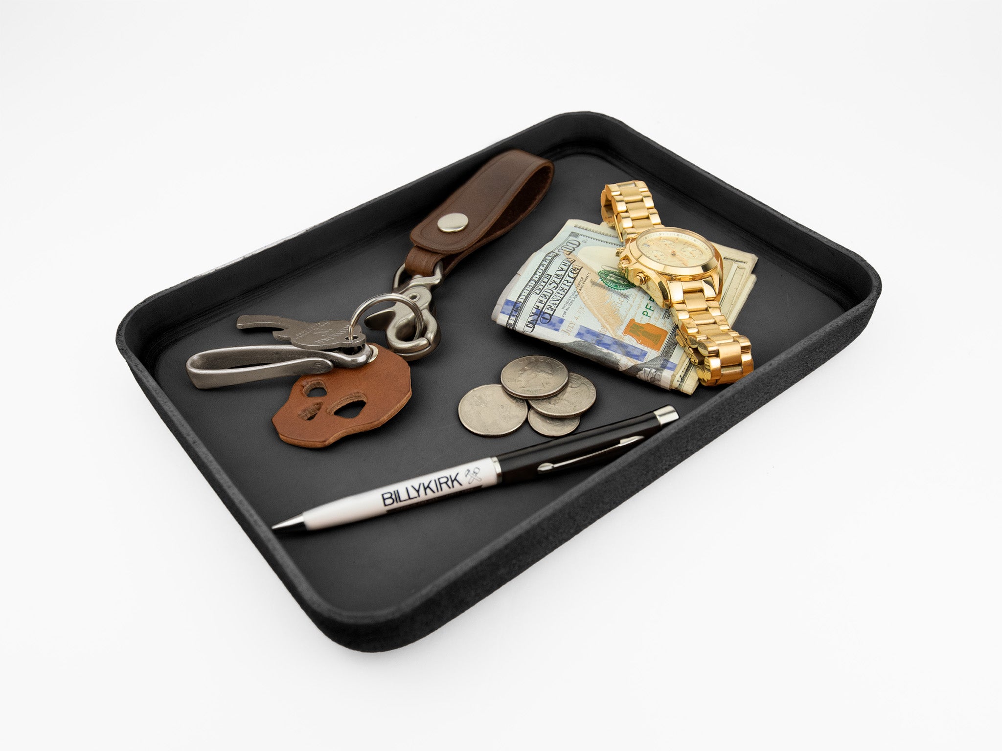 A black tray holding a set of keys with a leather keychain, a stack of cash, a gold wristwatch, some coins, and a pen labeled BILLYKIRK on a white background.