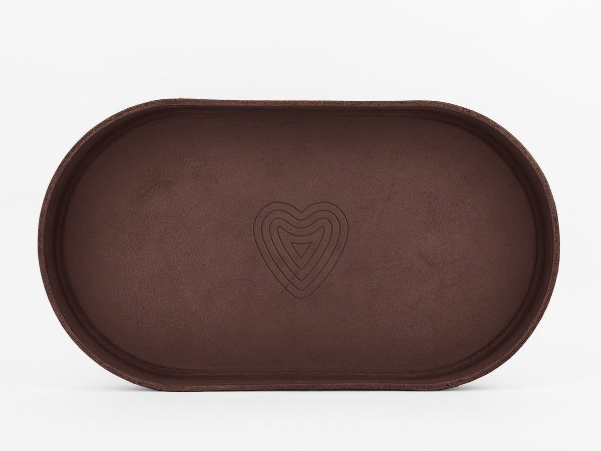 A brown, oval-shaped tray with a minimalist design and a concentric heart pattern embossed in the center, set against a plain white background.

