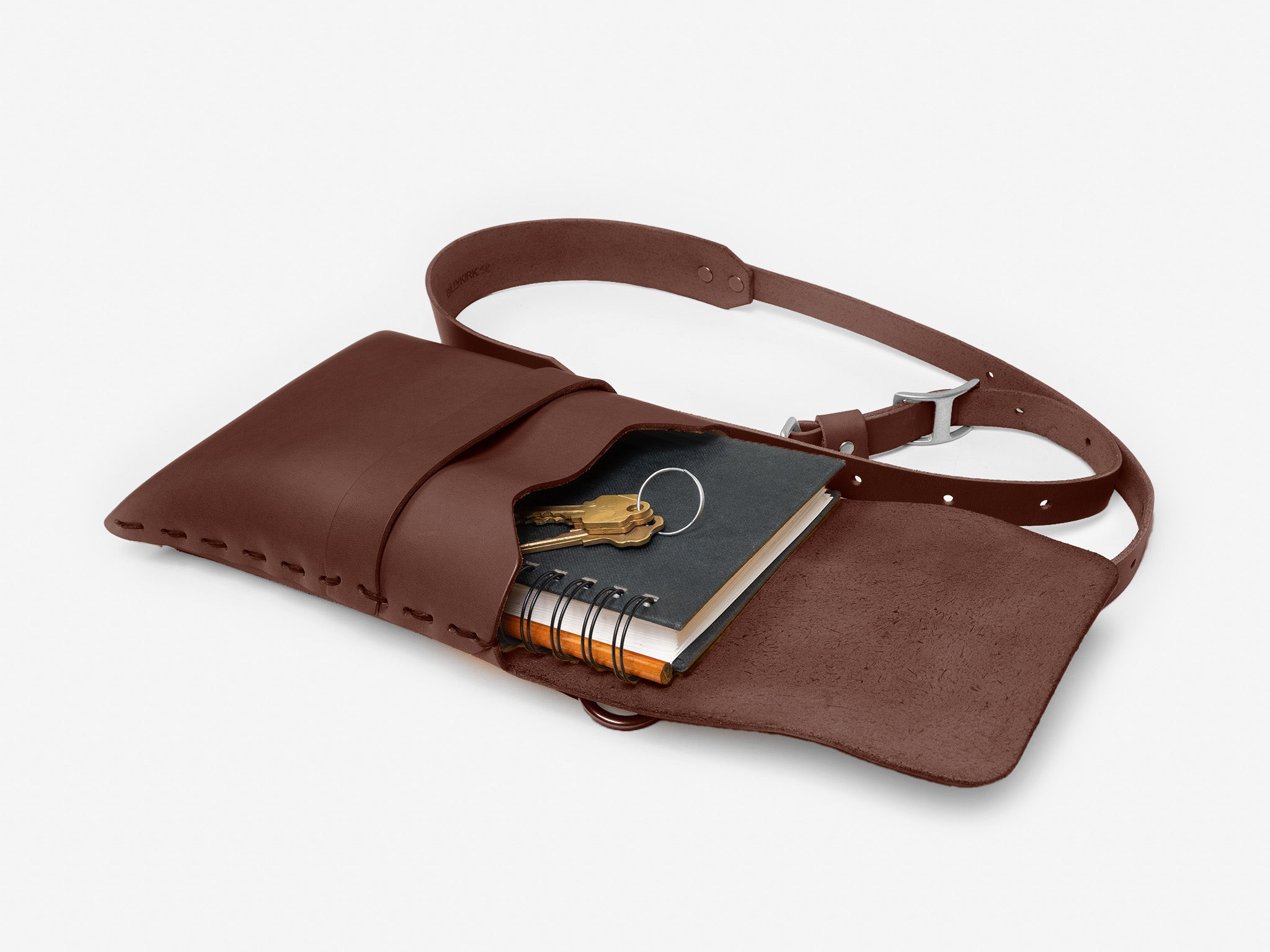 A brown leather belt bag lies open, revealing a spiral notebook and a set of two keys on a keyring inside, against a plain white background.
