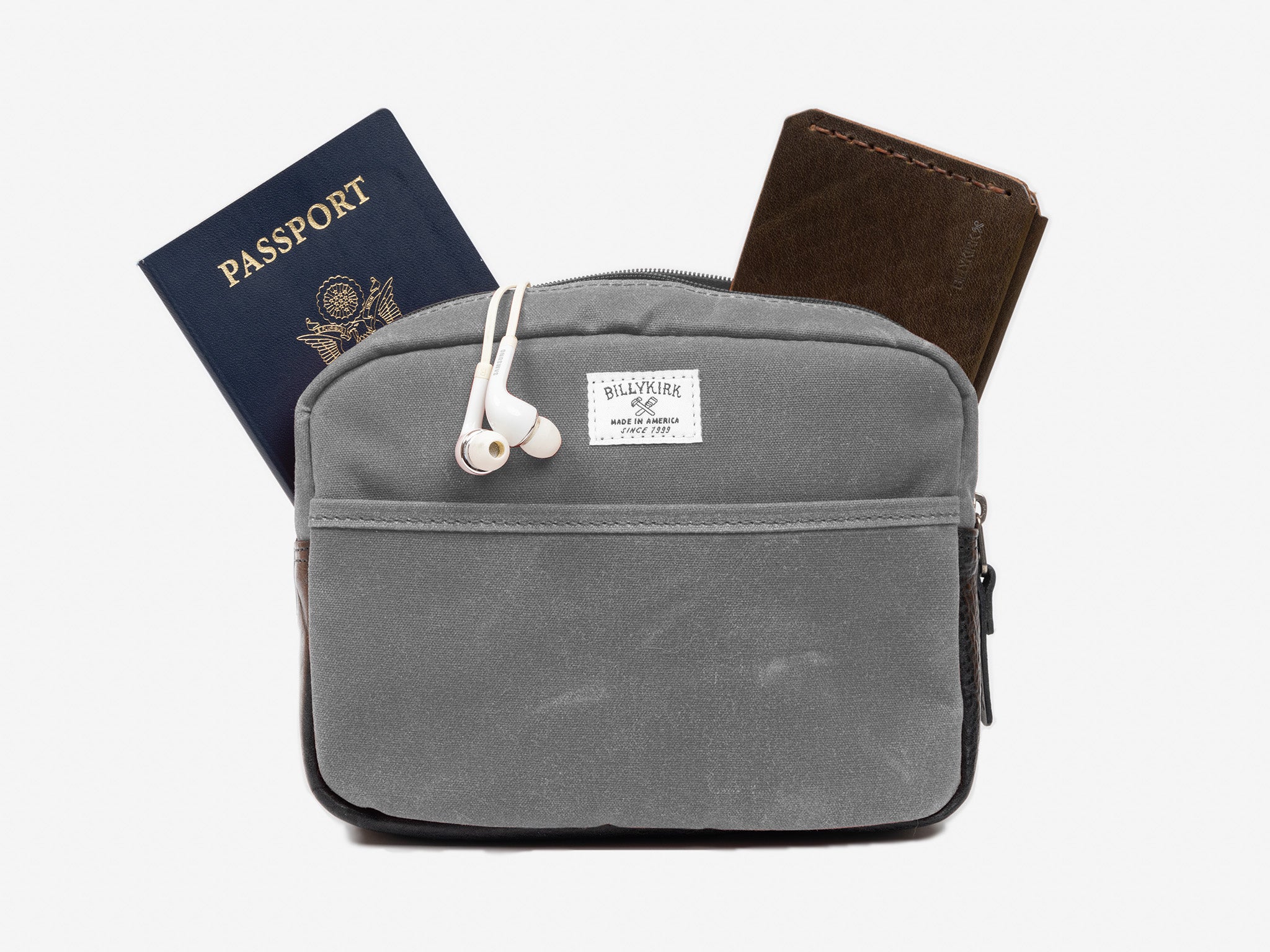A small ash waxed canvas crossbody bag with a wallet, passport, and white earphones sticking out of the zippered top. The bag has a front pocket, and a cream colored rectangular Billykirk brand patch stitched on the front. 