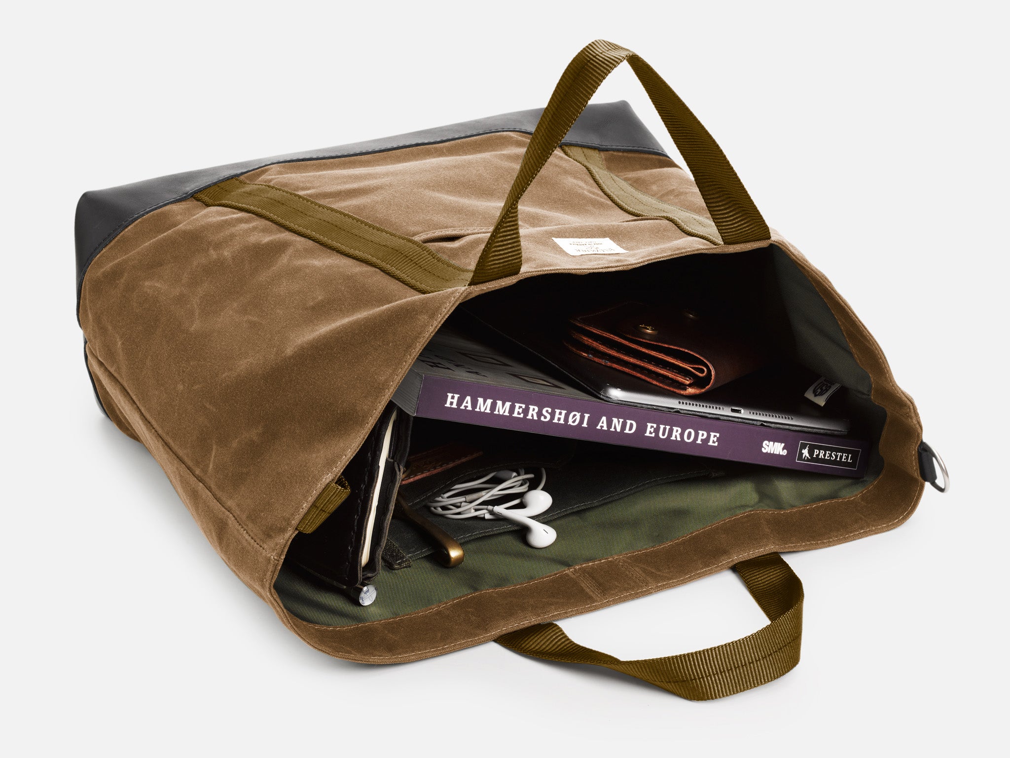 A brush brown tote bag, resting on its back, partially opened, reveals a book titled Hammershoi and Europe, a brown wallet, a tablet, and white earphones neatly inside.