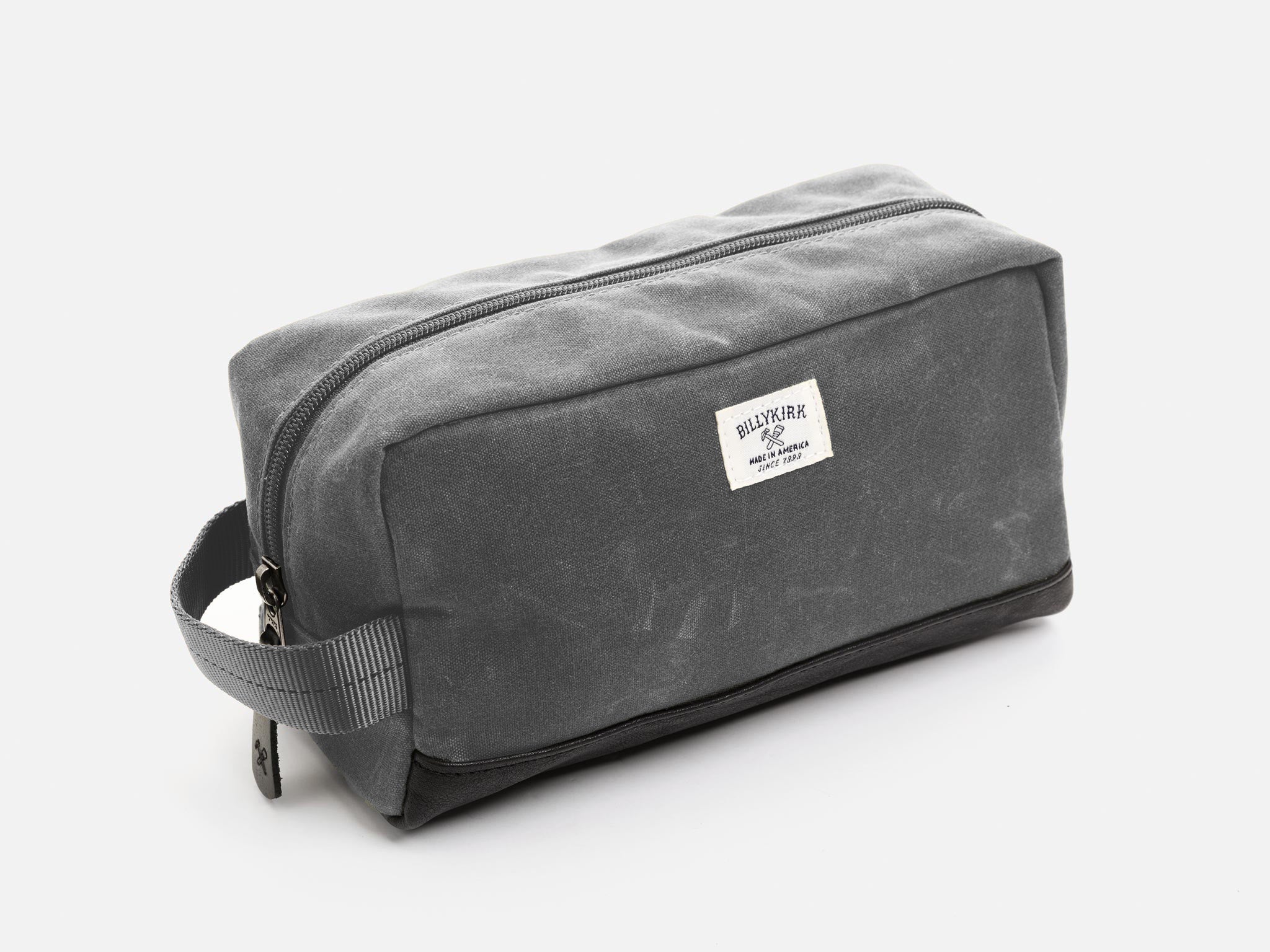 A rectangular ash grey waxed canvas toiletry bag with a zipper, a grey nylon webbing handle, a black leather base, and a off-white Billykirk brand label on the front.