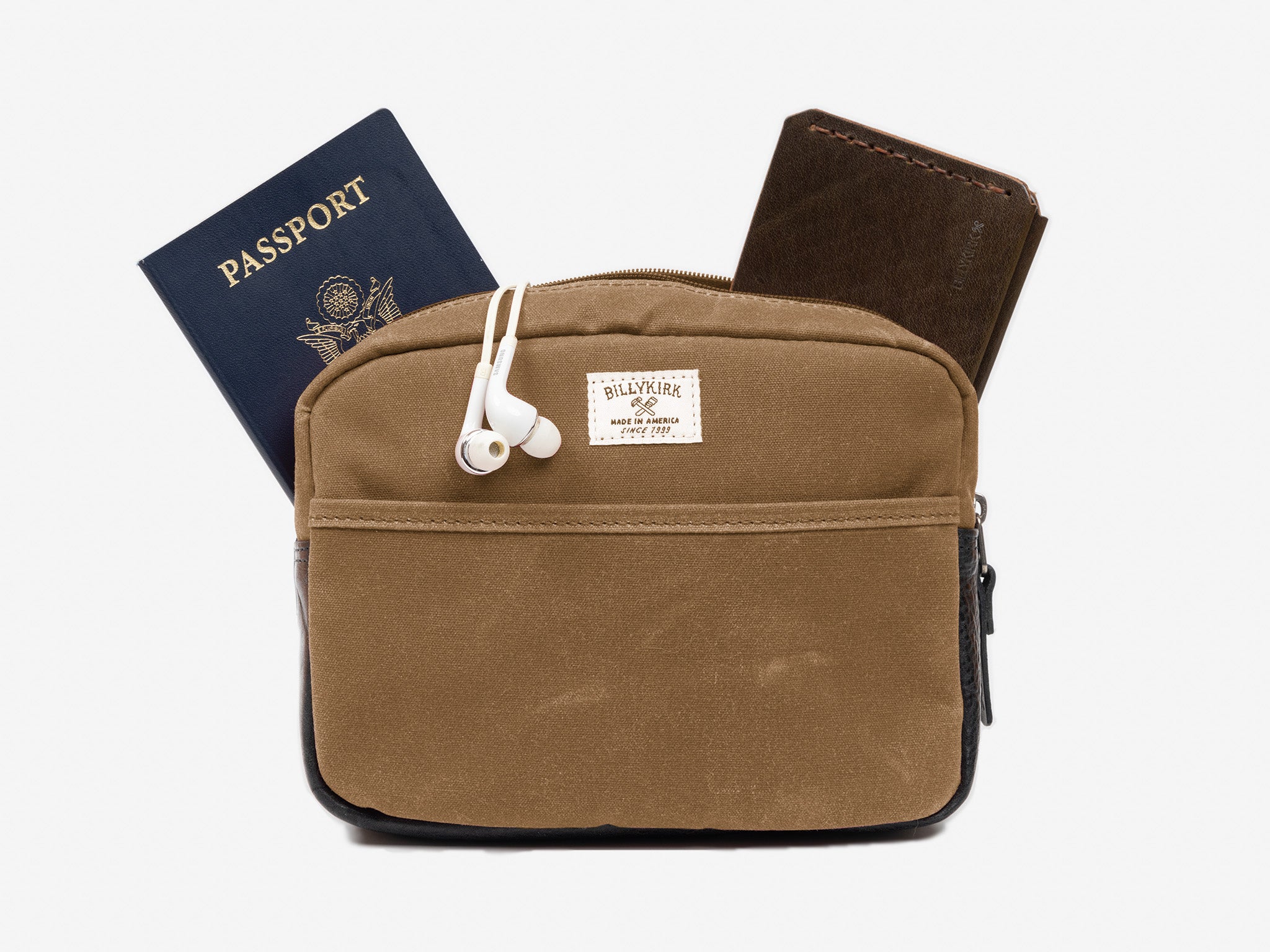 A small brush brown waxed canvas crossbody bag with a wallet, passport, and white earphones sticking out of the zippered top. The bag has a front pocket, and a cream colored rectangular Billykirk brand patch stitched on the front. 
