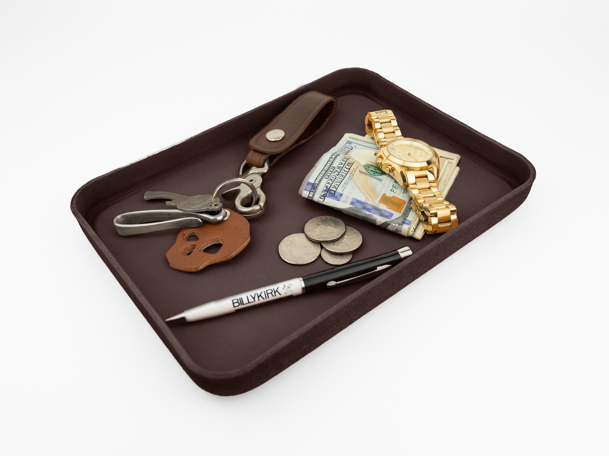 A brown tray holding a set of keys with a leather keychain, a stack of cash, a few coins, a gold wristwatch, and a black and white pen labeled BILLYKIRK on a white background.