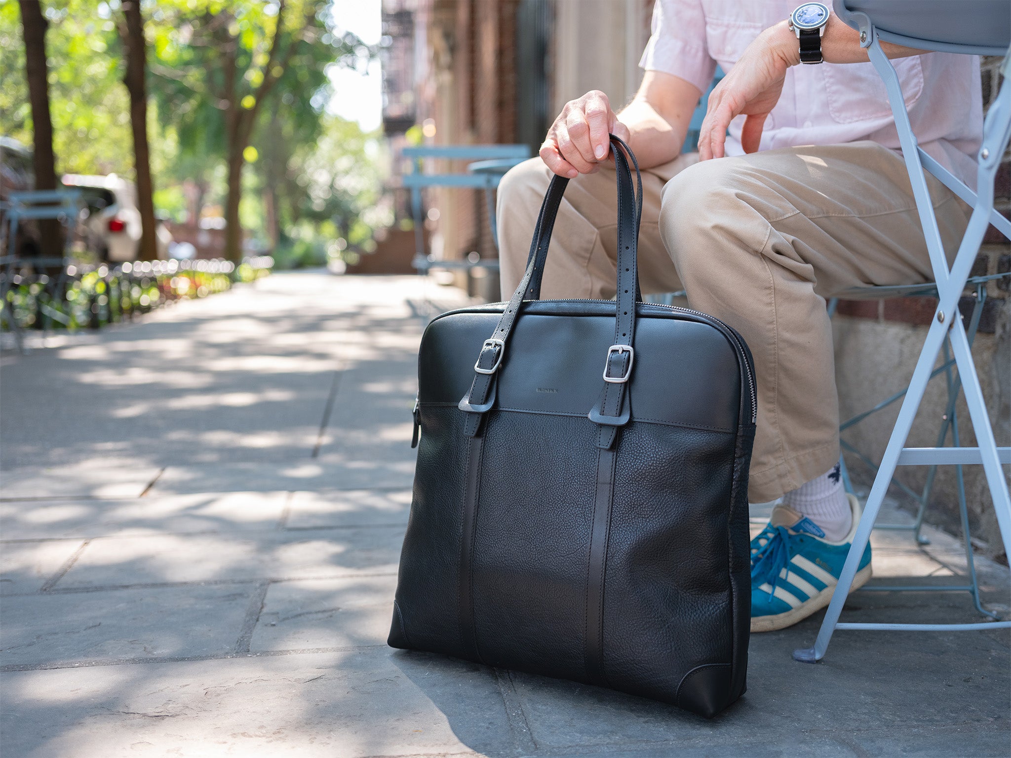A person sitting on a folding chair outdoors holds the handle of a large black leather briefcase resting on the pavement. The person wears khaki pants, blue-and-white sneakers, and a watch. Trees and benches line the sidewalk.