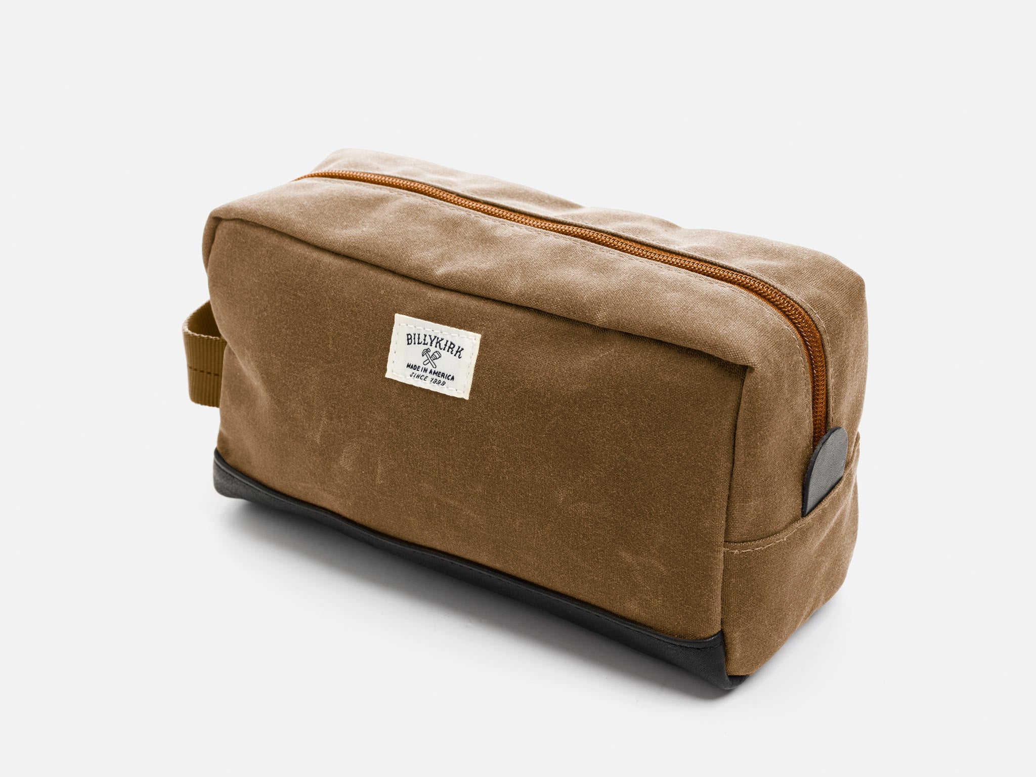 A rectangular brush brown waxed canvas toiletry bag with a zipper, a brush brown nylon webbing handle, a black leather base, a leather tab to help close the zipper, and an off-white Billykirk brand label on the front.