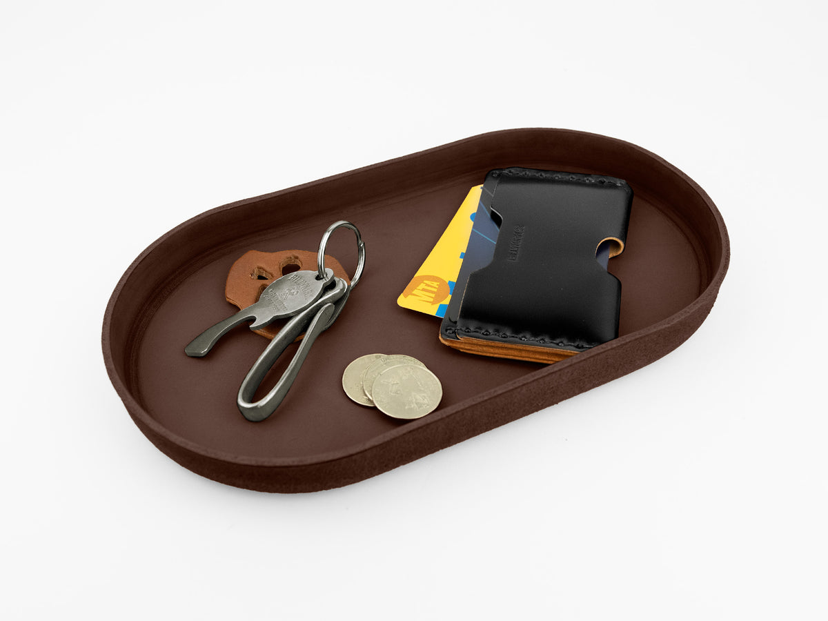 A brown leather tray holding a keychain with keys, a few coins, a black leather wallet, and some yellow and blue cards, all placed on a white background.