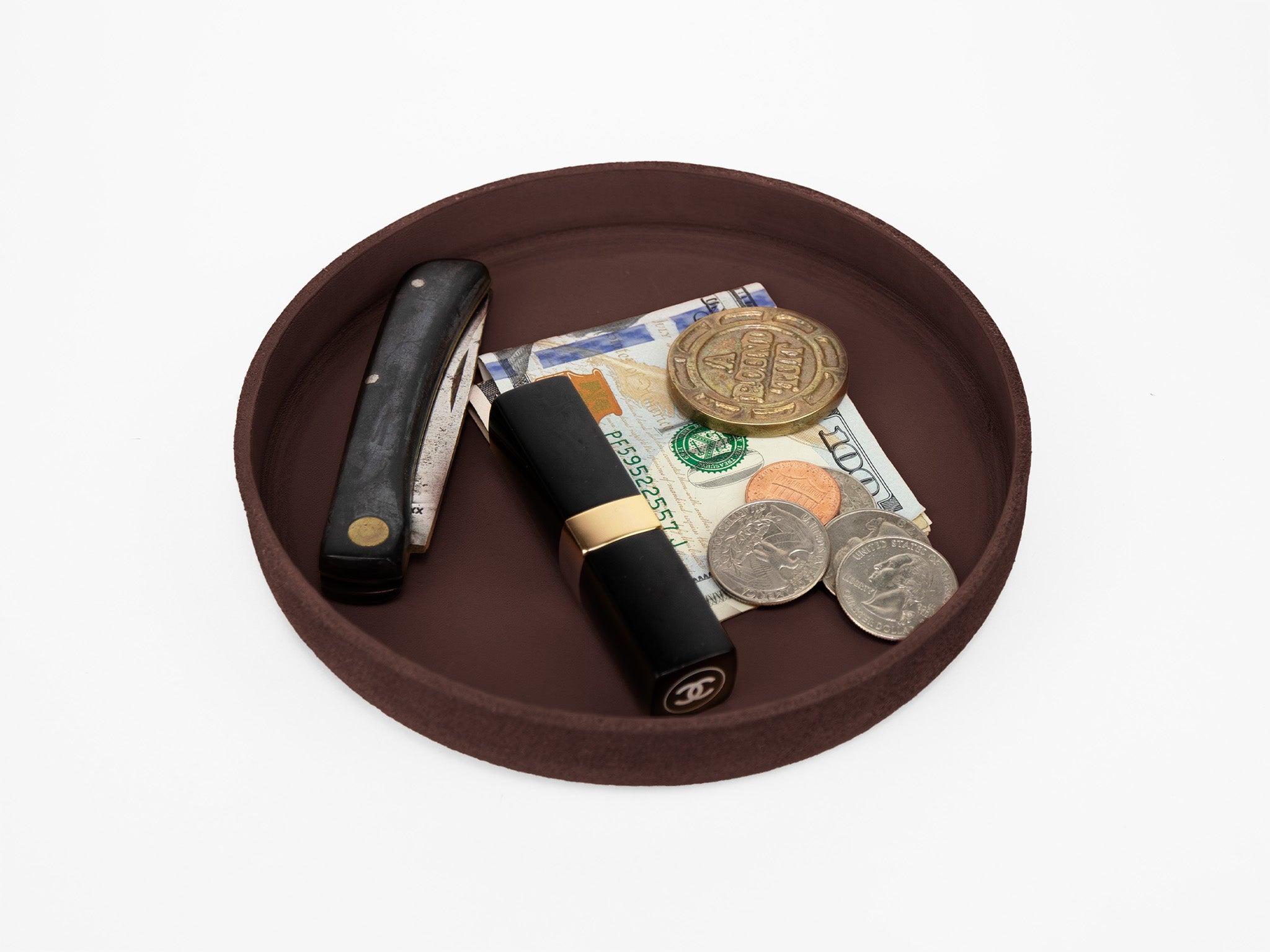 A round brown tray holds a straight razor, folded U.S. dollar bills, a gold coin, three silver coins, and a black and gold Chanel lipstick on a white background.