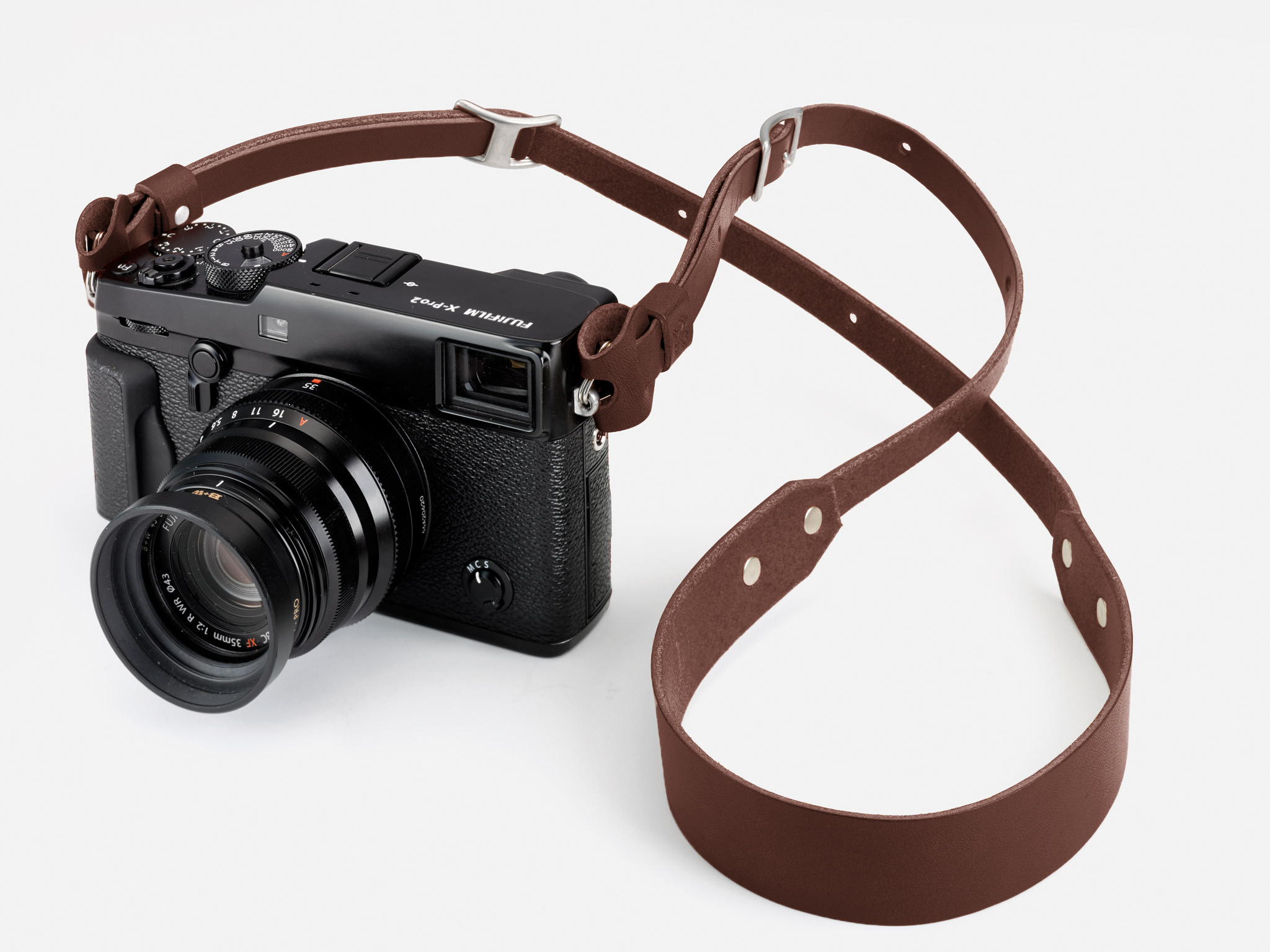 A Fujifilm X-Pro2 digital camera with a brown leather strap is placed on a white surface. The camera lens is facing forward and the strap is loosely arranged around it.