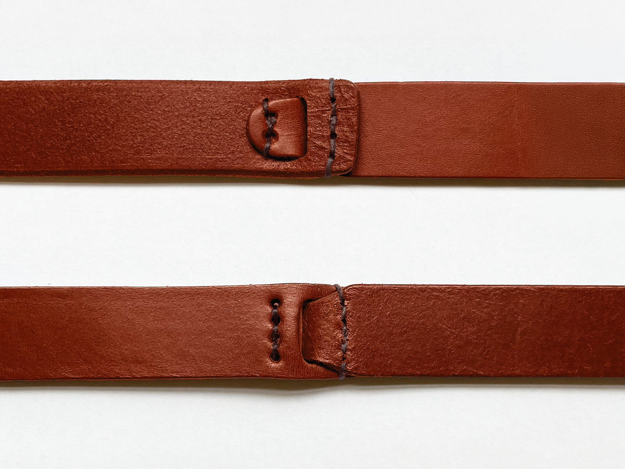 Two brown leather straps laid out parallel on a white background, each featuring stitched rectangular ends with holes and a slot for fastening.