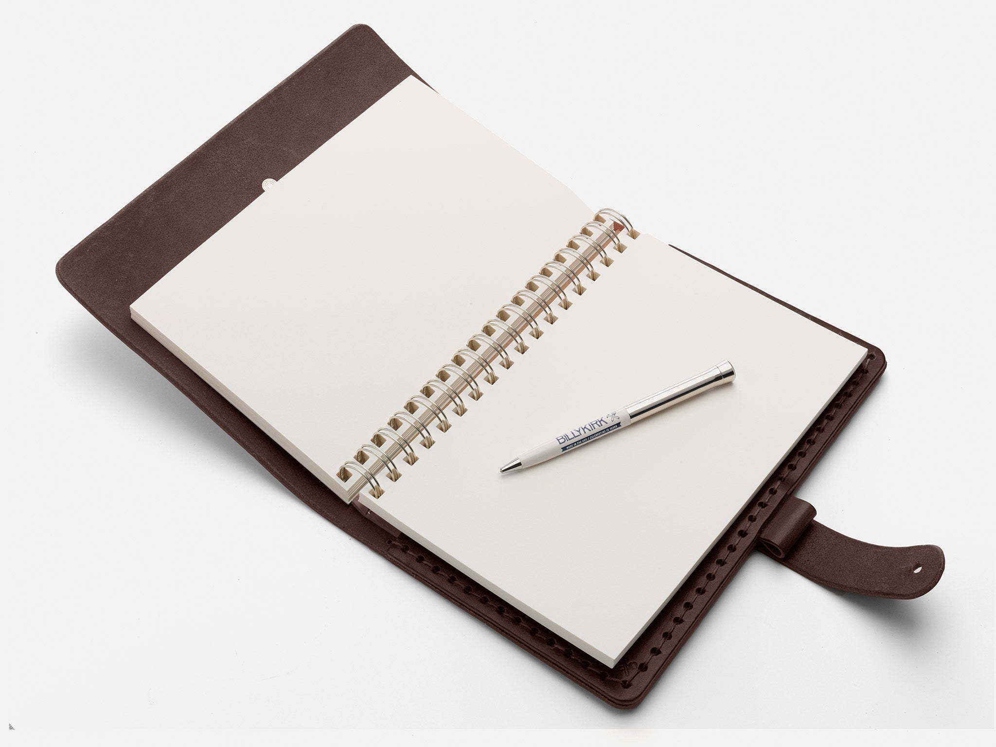 An open brown leather notebook with blank spiral-bound pages, lying on a white surface. A silver pen is placed diagonally across the pages.