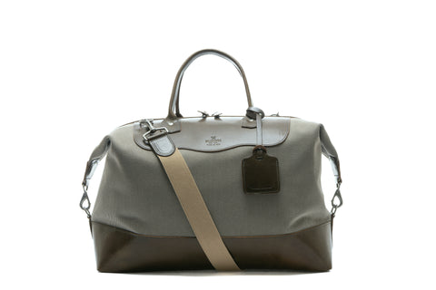 Taupe colored duffel bag with olive leather and a strap and luggage tag on a white background